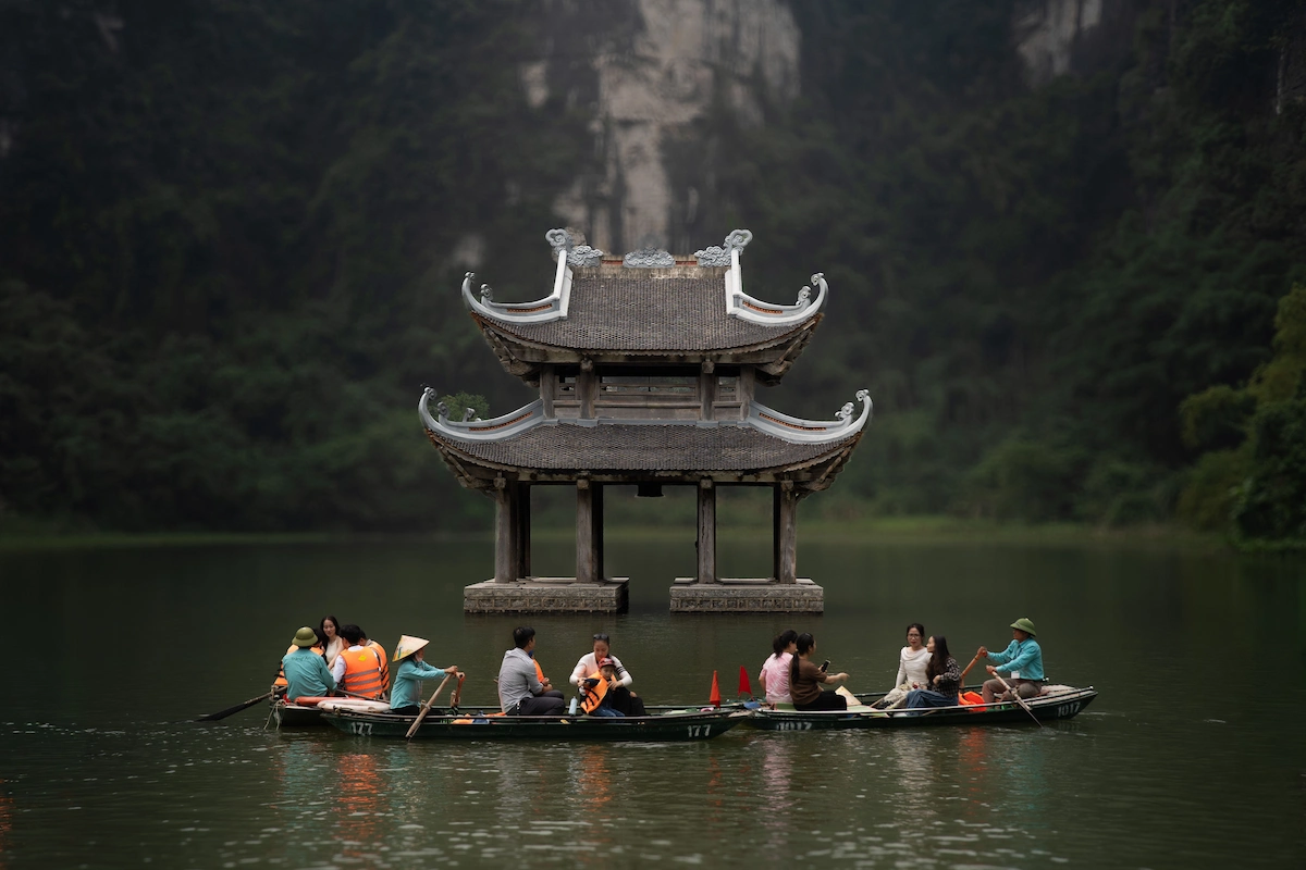 Tourists taking a Trang An Ninh Binh boat tour past a traditional pavilion on the water